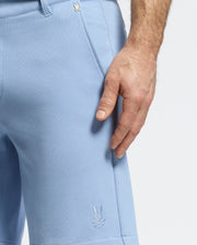 A person models Psycho Bunny's MENS SHIRO KNIT HONEYCOMB SHORT (B6R585F200) in light blue with a honeycomb pattern and small embroidered logo near the hem, resting a hand by the versatile shorts against a plain white background.