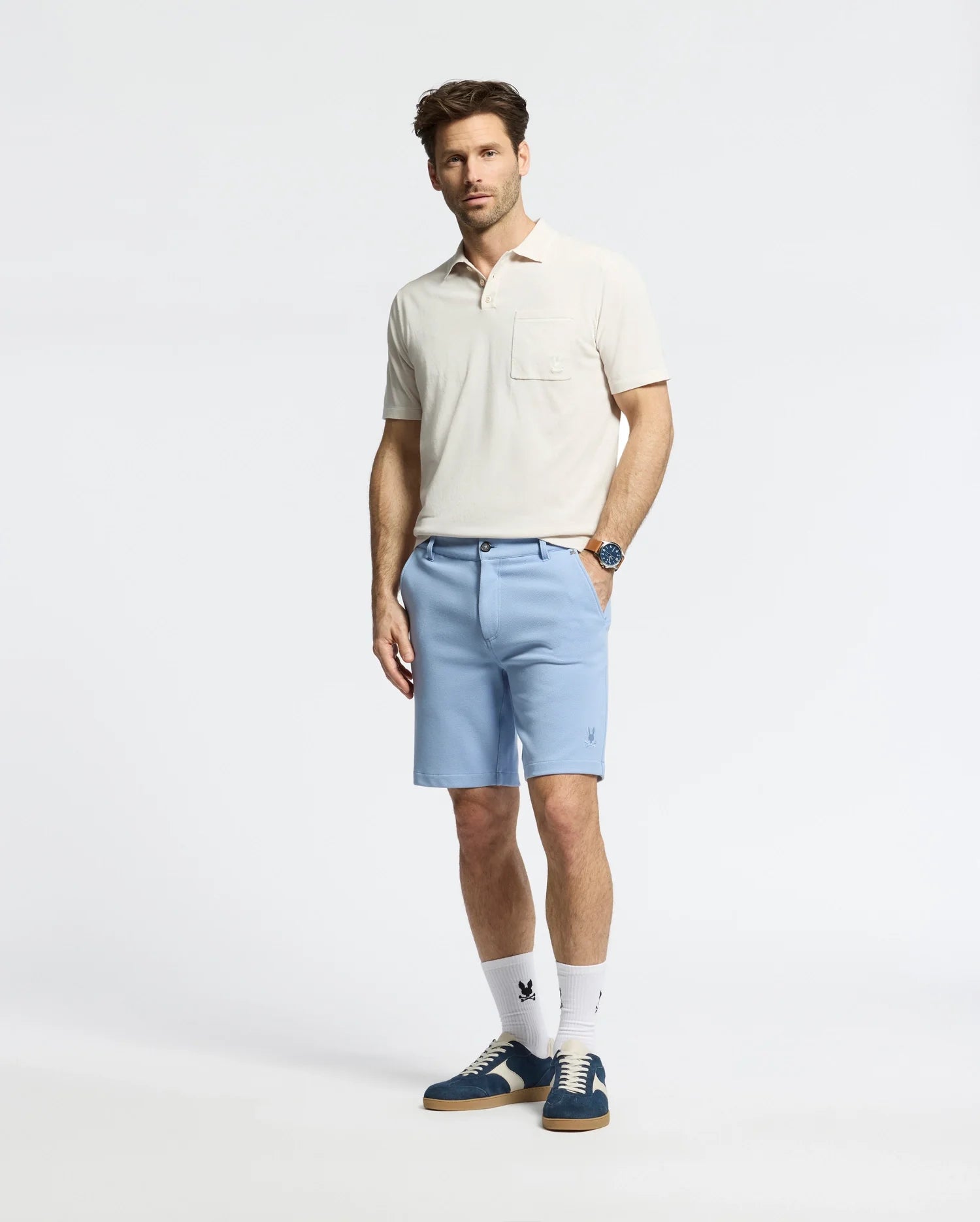 A dark-haired man stands against a plain background in light beige piqué with honeycomb texture, paired with light blue MENS SHIRO KNIT HONEYCOMB SHORT by Psycho Bunny. He's accessorized with a watch, white socks, and blue sneakers featuring a white leaf logo.