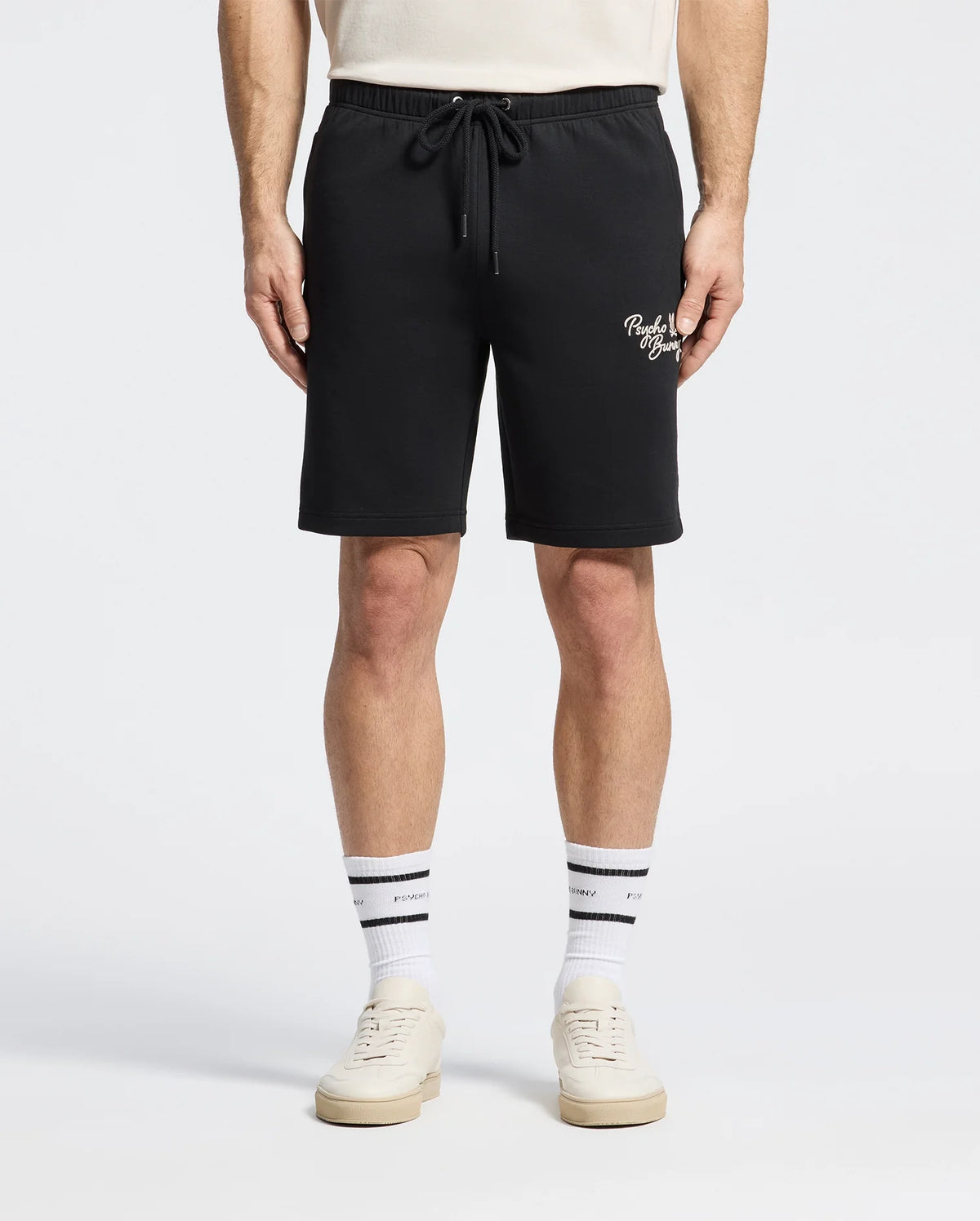 A person in a white T-shirt models the Psycho Bunny MENS DESERT EMBROIDERED SWEATSHORT in black, featuring white text on the left. Paired with white sneakers and socks with a black stripe and text.