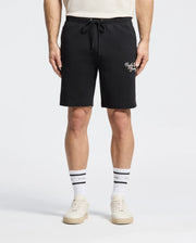 A person in a white T-shirt models the Psycho Bunny MENS DESERT EMBROIDERED SWEATSHORT in black, featuring white text on the left. Paired with white sneakers and socks with a black stripe and text.