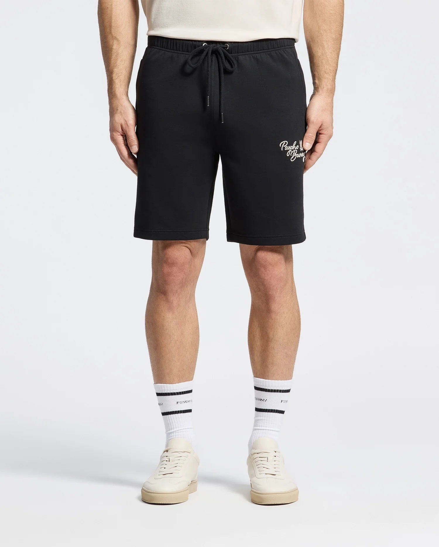 A person in a white T-shirt models the Psycho Bunny MENS DESERT EMBROIDERED SWEATSHORT in black, featuring white text on the left. Paired with white sneakers and socks with a black stripe and text.