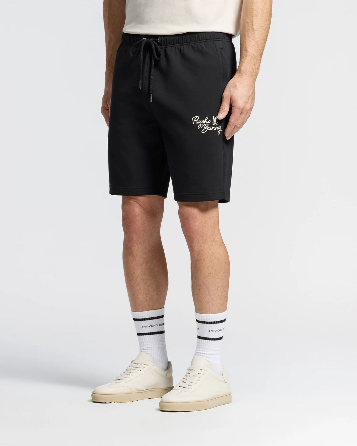 The individual is dressed in a white T-shirt, Psycho Bunny's MENS DESERT EMBROIDERED SWEATSHORT - B6R617F200 featuring white text and a leg logo, white socks with black stripes and text, and sleek white sneakers. The setting is a plain white backdrop.