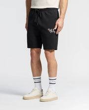 The individual is dressed in a white T-shirt, Psycho Bunny's MENS DESERT EMBROIDERED SWEATSHORT - B6R617F200 featuring white text and a leg logo, white socks with black stripes and text, and sleek white sneakers. The setting is a plain white backdrop.