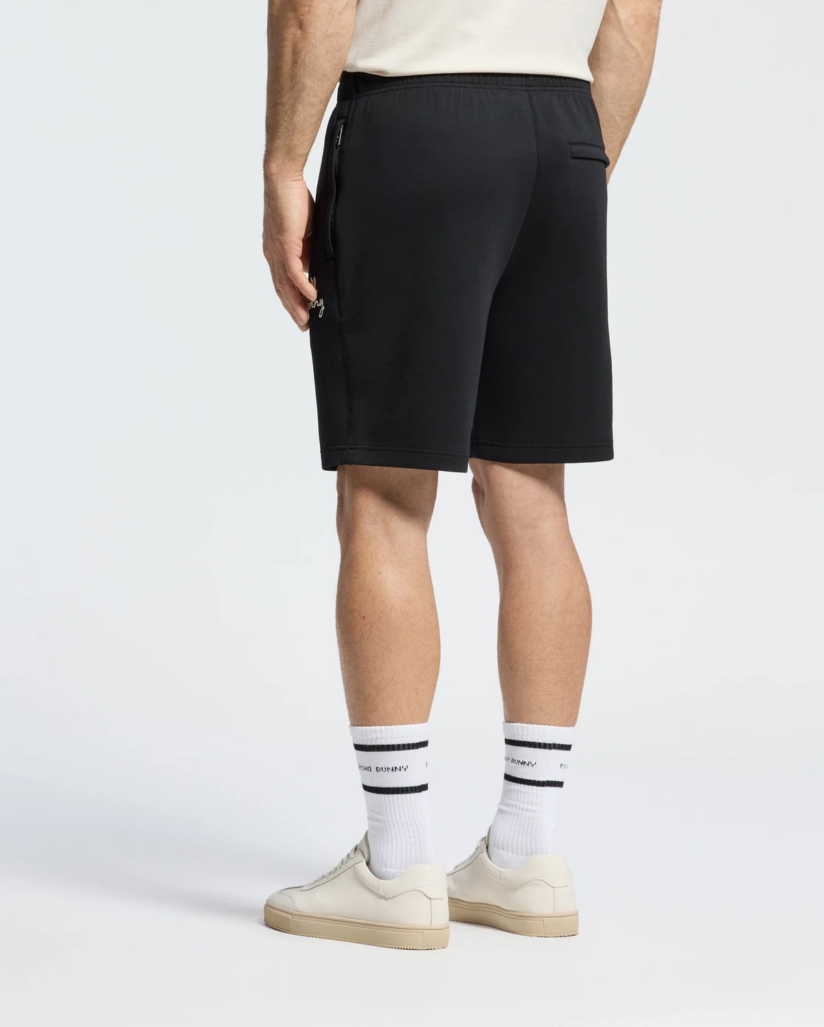 A back view shows a person in Psycho Bunny's MENS DESERT EMBROIDERED SWEATSHORT (B6R617F200), paired with moisture-wicking white socks featuring black stripes and white sneakers, all set against a plain white background to highlight the comfort of this casual outfit for an active lifestyle.