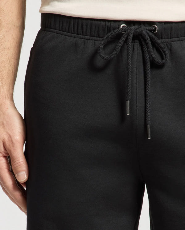 A close-up shows a person's waist wearing Psycho Bunny's MENS DESERT EMBROIDERED SWEATSHORT - B6R617F200 in black. The relaxed fit and metal-tipped drawstring are highlighted, with the left hand resting at the side. The soft modal-blend fabric stands out against a plain, light background.