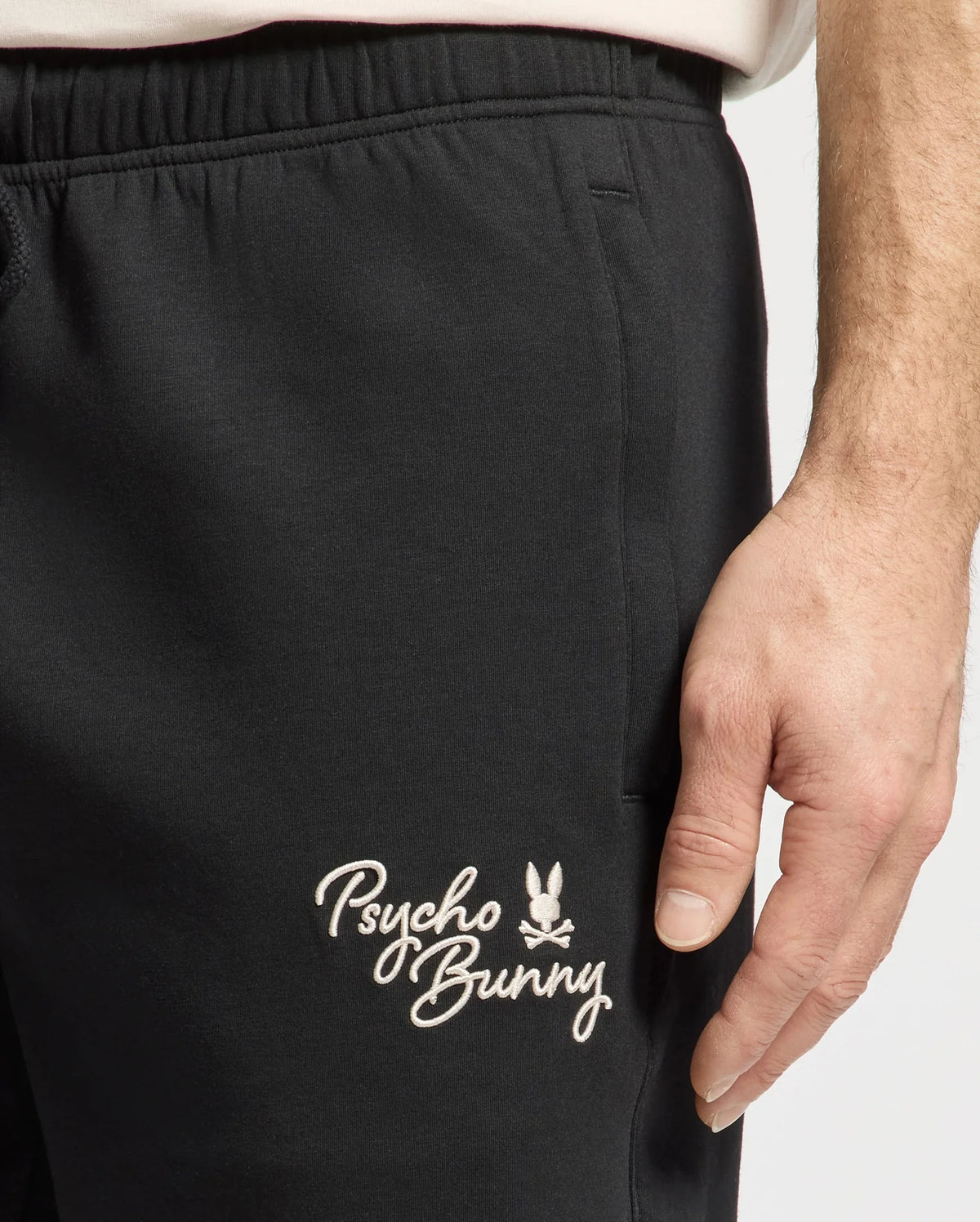 Close-up of a hand on a thigh, wearing MENS DESERT EMBROIDERED SWEATSHORT - B6R617F200 by Psycho Bunny, featuring their embroidered logo with a small bunny.