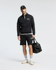 A man in a Psycho Bunny Desert Embroidered Sweatshort holds a black bag. He's wearing a black cap, white sneakers, socks with black stripes, and a moisture-wicking sweatshirt. The shorts and sweatshirt feature matching white logos against a plain background.