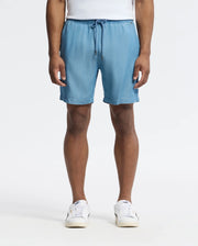 A person wears Psycho Bunny's MENS KERVIN LIGHTWEIGHT DENIM WOVEN SHORT - B6R633F200 in light blue, featuring an elasticized waist and drawstring, paired with white sneakers and standing against a plain white background, hands resting by their sides.