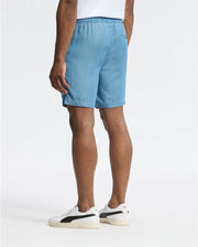 Someone wears the MENS KERVIN LIGHTWEIGHT DENIM WOVEN SHORT - B6R633F200 from Psycho Bunny with embroidered bunnies and white sneakers featuring black accents. The image shows their legs and part of their torso from the back against a plain white background.