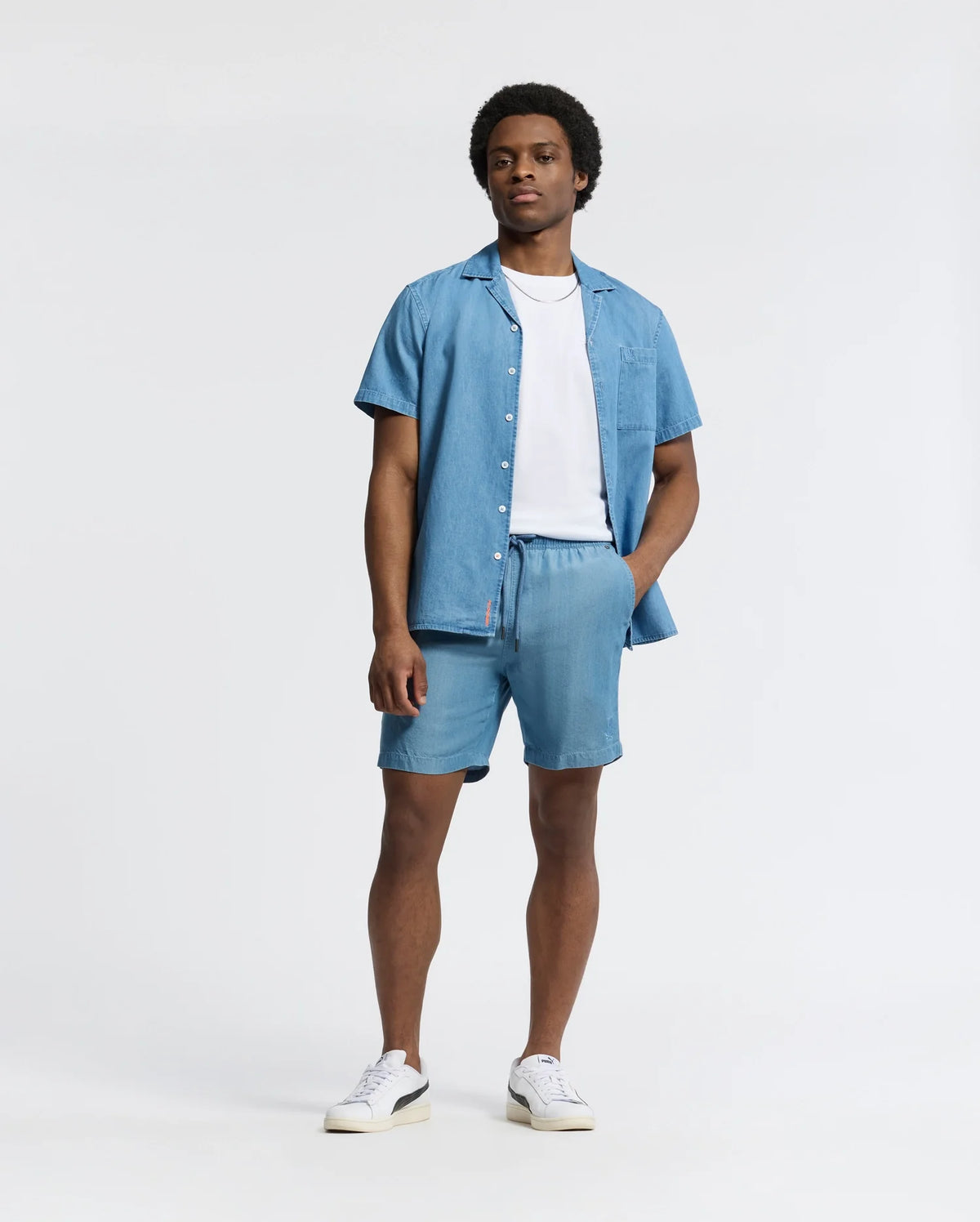 A person stands wearing a light blue short-sleeve button-up shirt over a white tee with Psycho Bunny's MENS KERVIN LIGHTWEIGHT DENIM WOVEN SHORT, featuring an elasticized waist. White sneakers finish the look against a plain white backdrop.