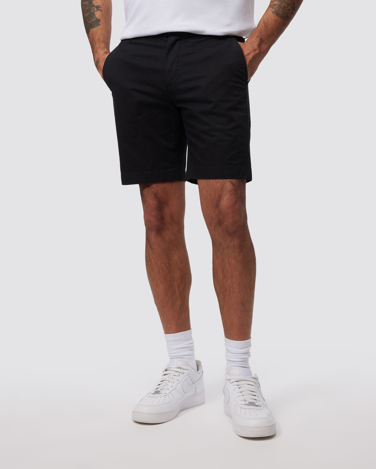 A person is shown from the waist down wearing a white T-shirt, slim fit black Psycho Bunny MENS HAYDEN SHORT - B6R757ARCE, and white sneakers. Their hands are in their pockets, and they are standing against a plain white background. The individual has tattoos on their forearms and is wearing breathable cotton white socks.