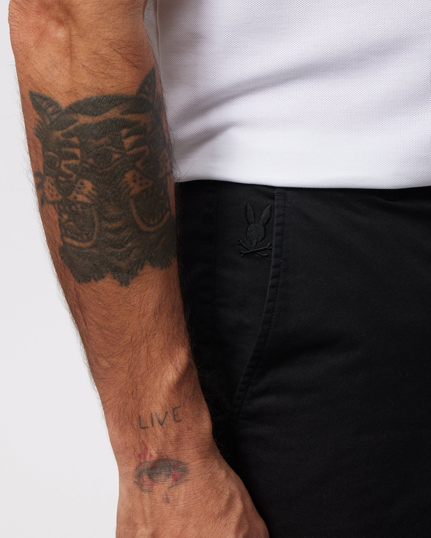 A person's arm with a large tattoo of a cat face is shown alongside Psycho Bunny MENS HAYDEN SHORT - B6R757ARCE with a small embroidered logo of a rabbit near the pocket. The person is wearing a white shirt made of breathable cotton. Their wrist has another tattoo that reads "LIVE" above a small eye design.
