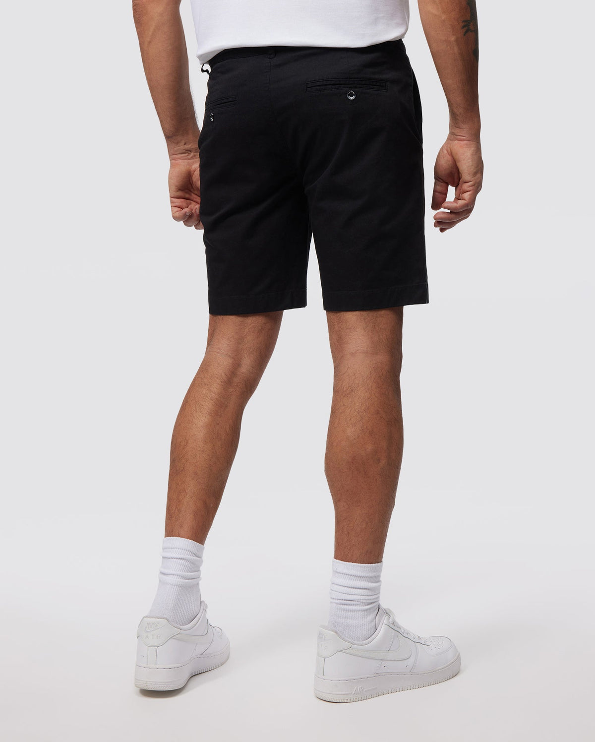 A person is standing facing away from the camera, wearing a white t-shirt, Psycho Bunny MENS HAYDEN SHORT - B6R757ARCE in black, white socks, and white sneakers. The slim fit ensemble is crafted from breathable cotton. The background is plain and neutral.