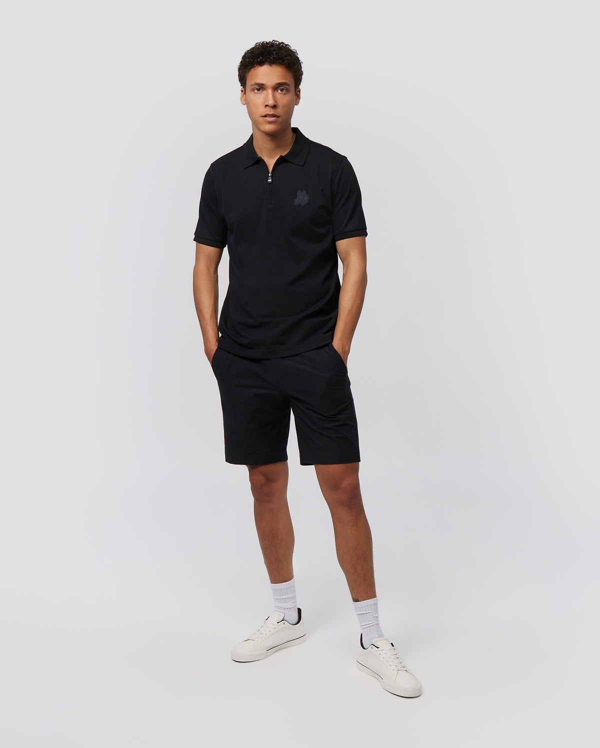 A man stands against a plain white background wearing a black polo shirt and Psycho Bunny MENS GABLE REGULAR FIT SPORT SHORT - B6R761ARCN. He has short, curly hair and is looking directly at the camera with his hands in his hidden zip pocket. He is also wearing white socks and white sneakers.