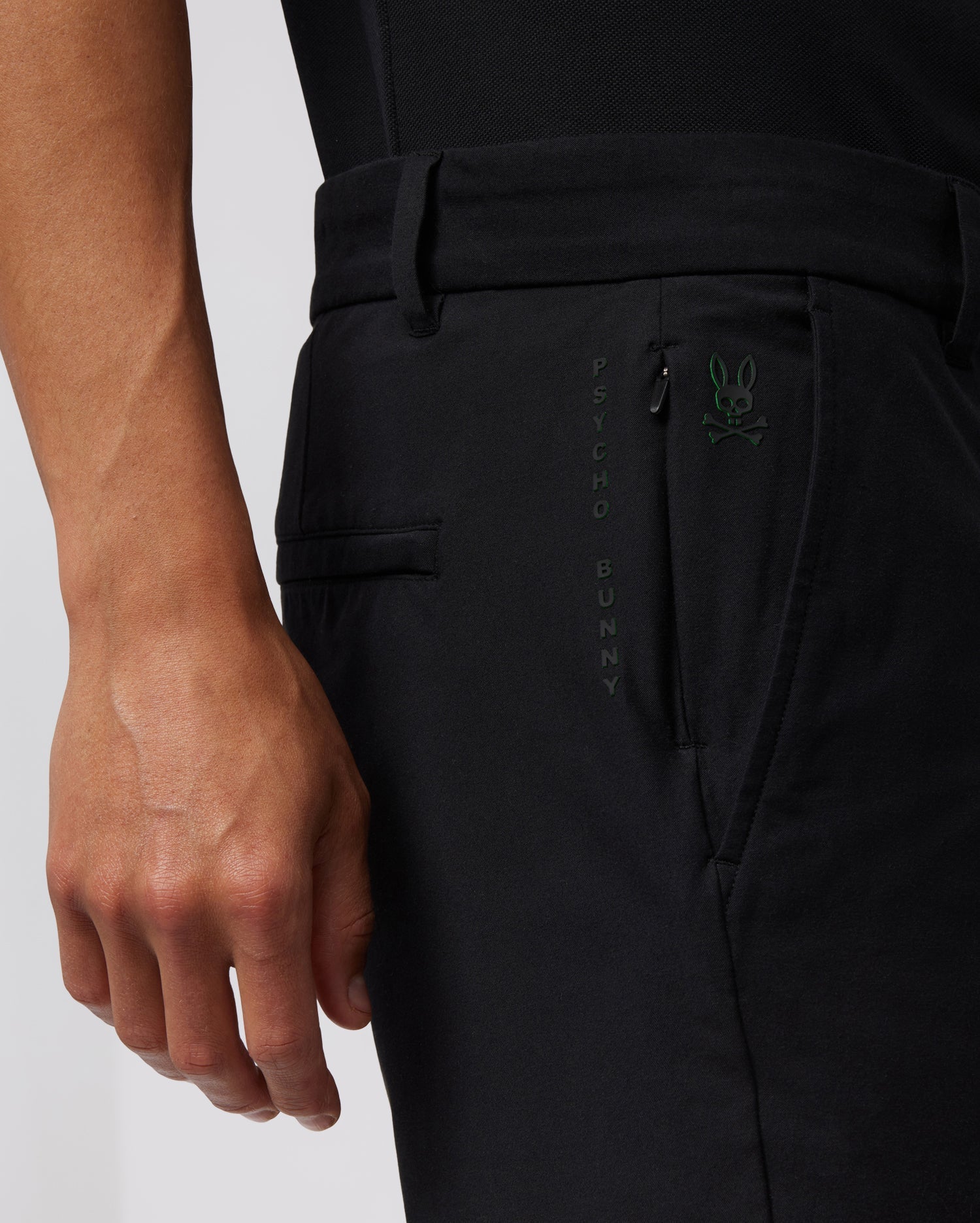 Close-up of a person wearing black pants with a small green Psycho Bunny logo and the words "PSYCHO BUNNY" vertically stitched near the pocket. The durable stretch cotton-nylon blend ensures comfort, while the person’s hand is visible resting alongside the hidden zip pocket. The background is plain and white. They are wearing MENS GABLE REGULAR FIT SPORT SHORT - B6R761ARCN by Psycho Bunny.