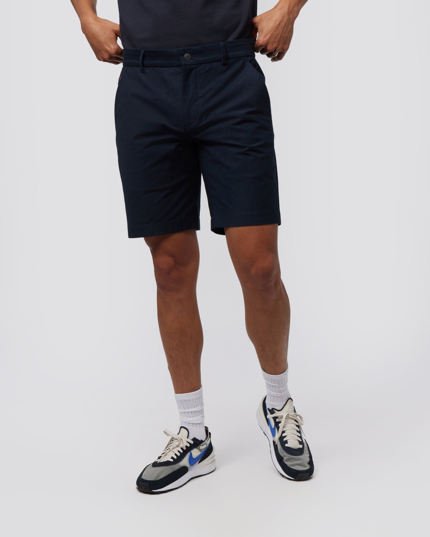 A person wearing knee-length, navy blue Psycho Bunny MENS GABLE REGULAR FIT SPORT SHORT - B6R761ARCN made from durable stretch cotton-nylon, white socks, and white sneakers with blue, black, and beige accents stands against a plain white background. The individual's hands are tucked into the pockets of their shorts.