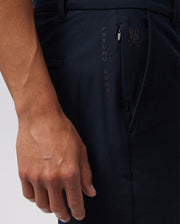 Close-up of a person wearing the MENS GABLE REGULAR FIT SPORT SHORT - B6R761ARCN by Psycho Bunny in dark-colored durable stretch cotton-nylon with the words "PSYCHO BUNNY" embroidered vertically next to the hidden zip pocket. The person's hand is relaxed by their side, partially revealing the right arm and a small logo of a bunny with crossbones.