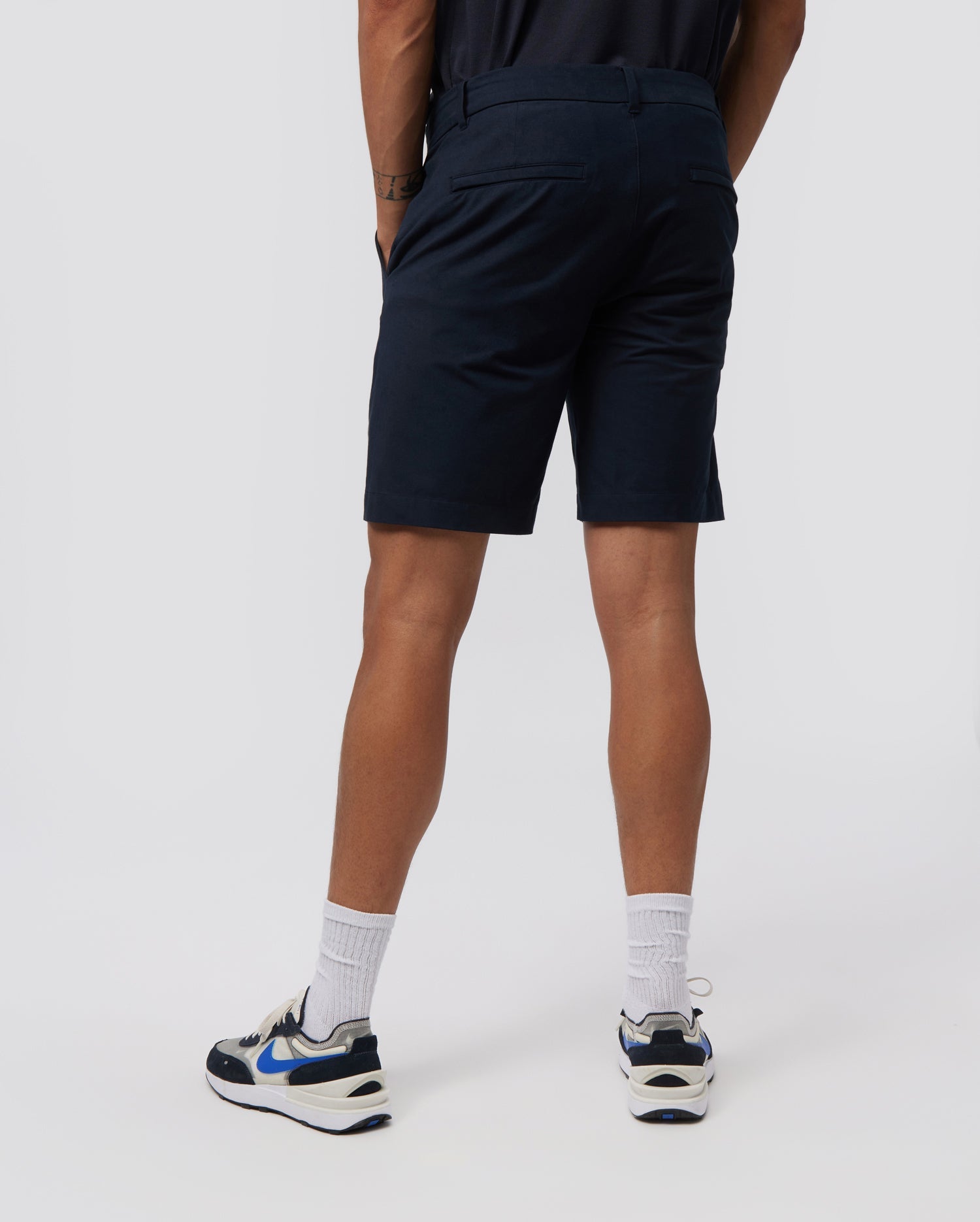 A person standing with their back to the camera, wearing Psycho Bunny MENS GABLE REGULAR FIT SPORT SHORT - B6R761ARCN made from durable stretch cotton-nylon, white socks, and white sneakers with blue and black accents. The background is a plain light gray.
