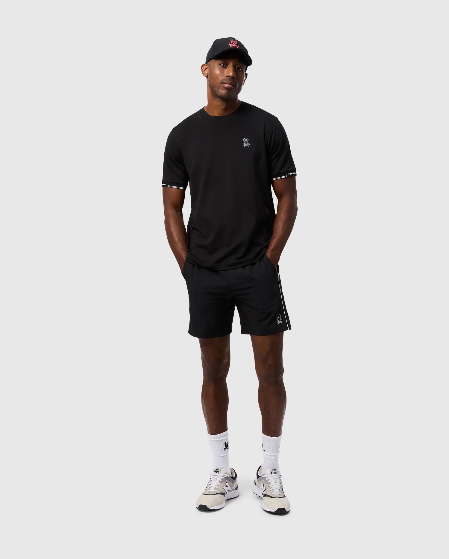 A person wearing a black T-shirt, polyester sport shorts from Psycho Bunny (MENS MANSFIELD SPORT SHORT - B6R807D200), and a black cap stands against a plain background. They are also wearing white sneakers and white socks, with their hands in their pockets.