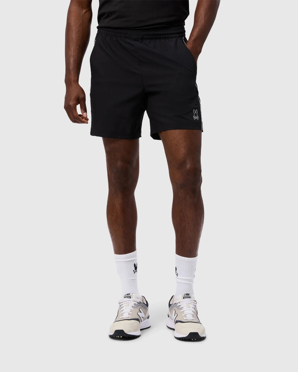 A person in a black t-shirt and Psycho Bunny MENS MANSFIELD SPORT SHORT - B6R807D200 is captured against a plain background. The black polyester athletic shorts feature a small white logo on the left leg. The look is completed with white socks and grey athletic shoes as the person is shown from shoulders down.
