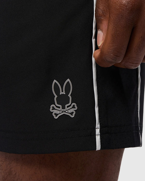 A close-up of a hand resting on the side of Psycho Bunny's MENS MANSFIELD SPORT SHORT - B6R807D200, crafted from breathable polyester. These regular fit black shorts showcase a small bunny head and crossbones logo along with white vertical stripes running down the side seam.