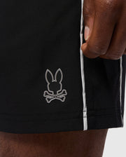 A close-up of a hand resting on the side of Psycho Bunny's MENS MANSFIELD SPORT SHORT - B6R807D200, crafted from breathable polyester. These regular fit black shorts showcase a small bunny head and crossbones logo along with white vertical stripes running down the side seam.