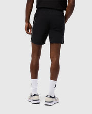 A person is viewed from the back wearing Psycho Bunny's MENS MANSFIELD SPORT SHORT - B6R807D200, which are regular-fit polyester shorts featuring a white stripe on the side against a plain light gray background. The outfit is complemented by white socks and running shoes.