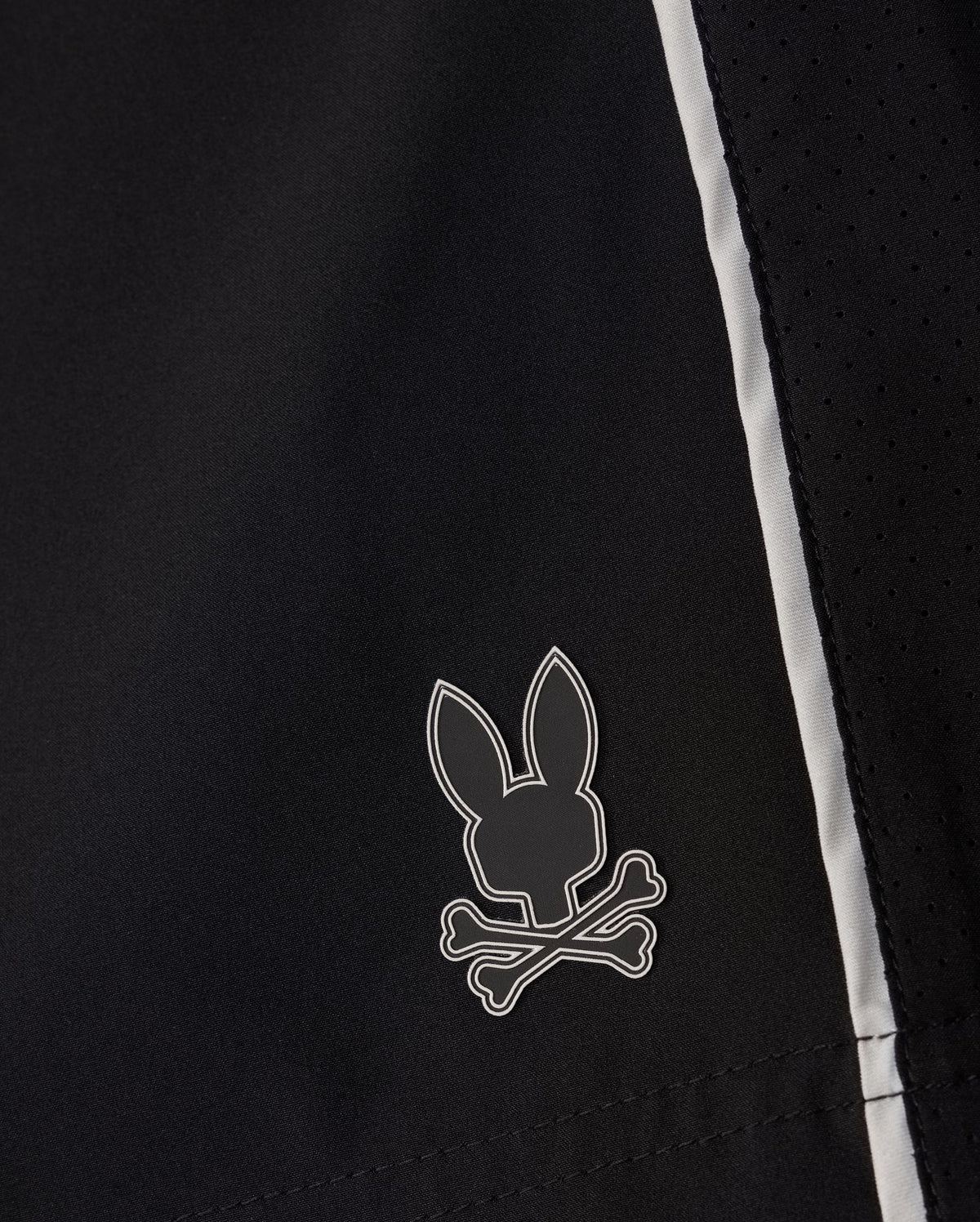 Close-up of the MENS MANSFIELD SPORT SHORT - B6R807D200 by Psycho Bunny in black polyester fabric, showcasing a small logo of a rabbit head above crossed bones. This whimsical take on the traditional skull and crossbones design is accentuated by its perforated texture and white accents on the side, providing a regular fit.