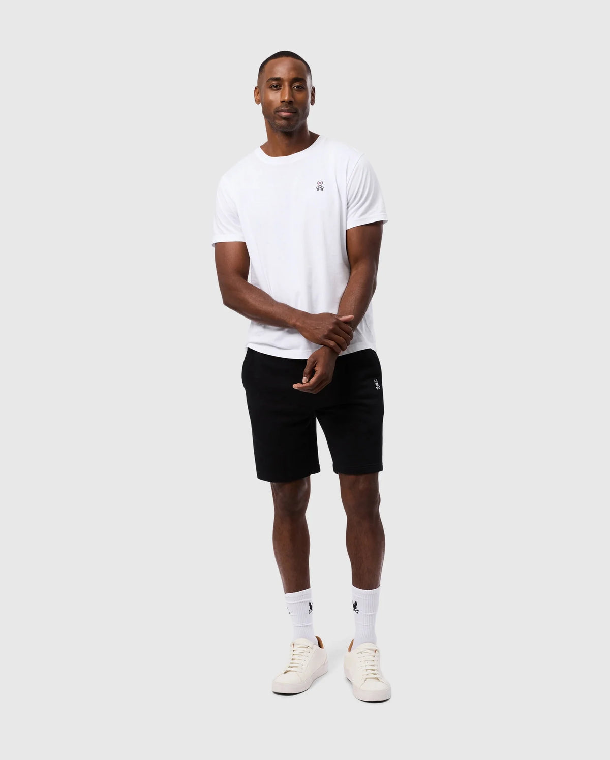 A man stands against a plain background, wearing a white t-shirt, Psycho Bunny's MENS LEVY FLEECE SWEATSHORT - B6R862D200, white socks, and white sneakers. His arms are relaxed at his sides, and he smiles gently towards the camera. The overall look is casual and sporty—an everyday staple for comfort and style.