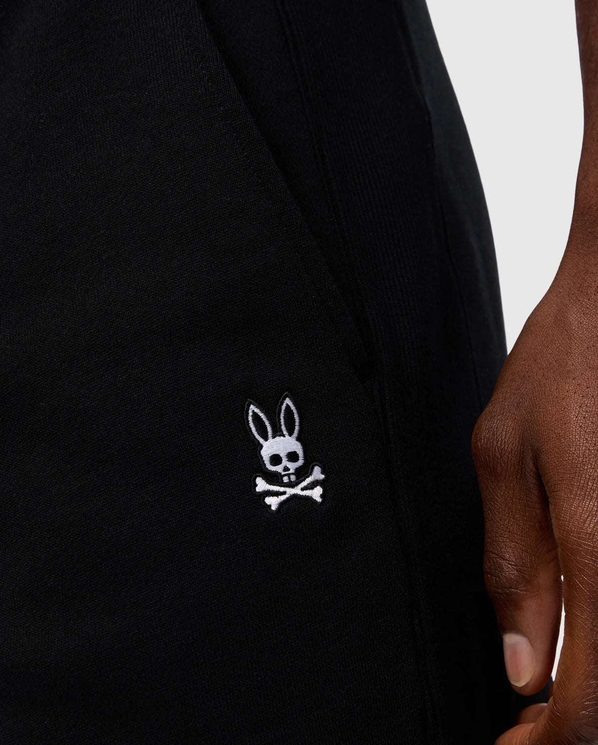 Close-up of a person holding Psycho Bunny's MENS LEVY FLEECE SWEATSHORT - B6R862D200 in black cotton, featuring an embroidered logo of a rabbit head with crossed bones below it, adding a playful twist to the classic skull and crossbones design.