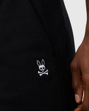 Close-up of a person holding Psycho Bunny's MENS LEVY FLEECE SWEATSHORT - B6R862D200 in black cotton, featuring an embroidered logo of a rabbit head with crossed bones below it, adding a playful twist to the classic skull and crossbones design.