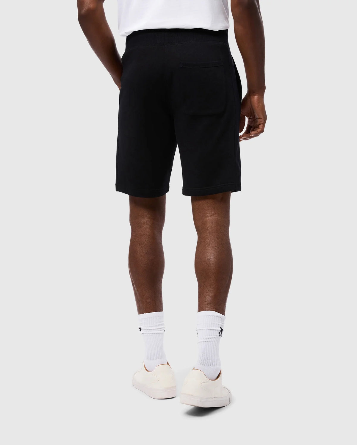 A person is standing with their back to the camera, wearing a white T-shirt, white socks, white sneakers, and Psycho Bunny's MENS LEVY FLEECE SWEATSHORT (B6R862D200). The background is plain and light-colored. The person's hands are relaxed by their sides.