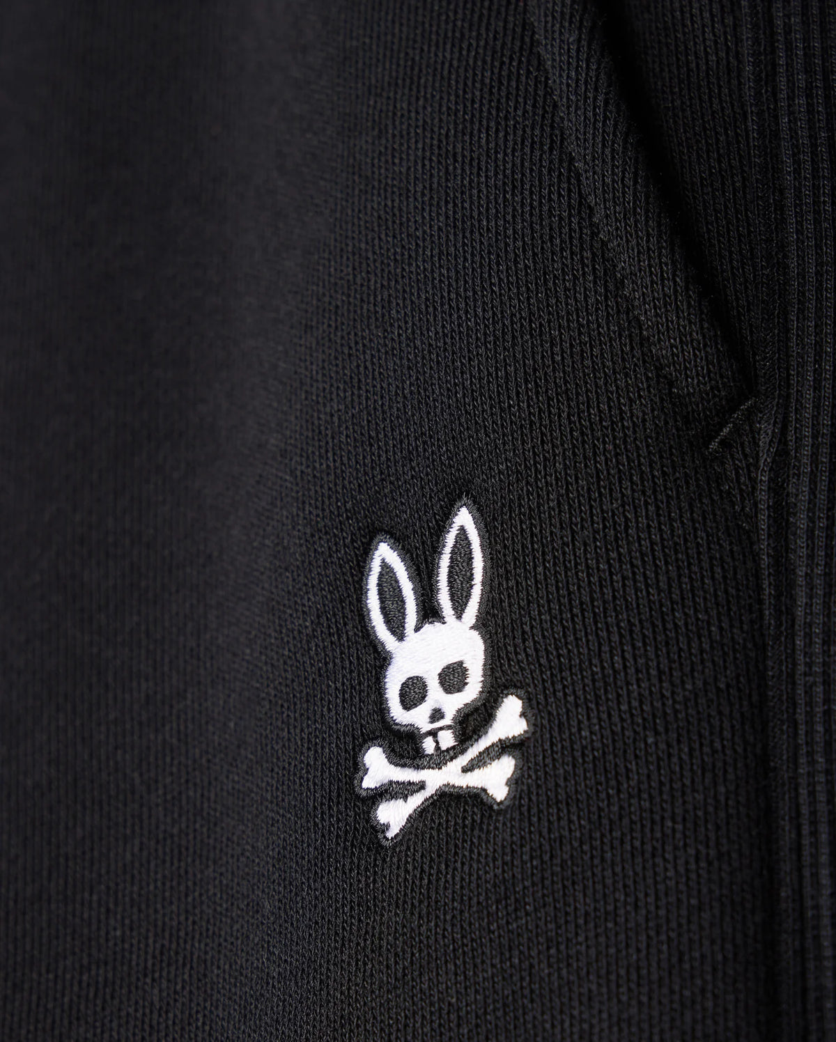 Close-up of the MENS LEVY FLEECE SWEATSHORT - B6R862D200 by Psycho Bunny, showcasing a black fabric garment adorned with an embroidered white logo featuring a rabbit head with long ears and a skull and crossbones beneath it. The texture of the cotton sweatshorts is visible, highlighting the detailed stitching of the emblem.