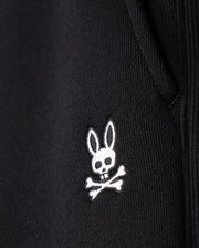 Close-up of the MENS LEVY FLEECE SWEATSHORT - B6R862D200 by Psycho Bunny, showcasing a black fabric garment adorned with an embroidered white logo featuring a rabbit head with long ears and a skull and crossbones beneath it. The texture of the cotton sweatshorts is visible, highlighting the detailed stitching of the emblem.