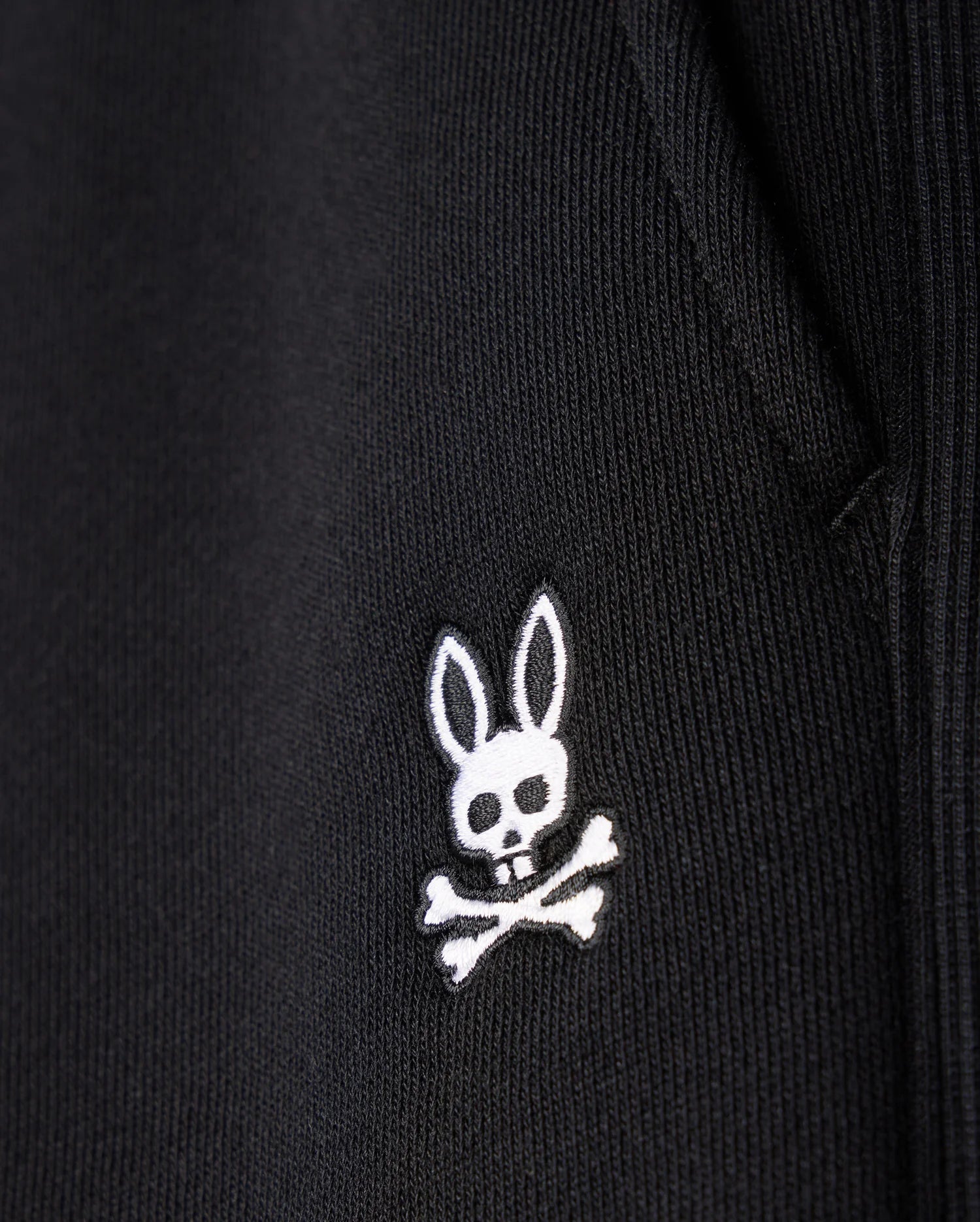 Close-up of the MENS LEVY FLEECE SWEATSHORT - B6R862D200 by Psycho Bunny, showcasing a black fabric garment adorned with an embroidered white logo featuring a rabbit head with long ears and a skull and crossbones beneath it. The texture of the cotton sweatshorts is visible, highlighting the detailed stitching of the emblem.
