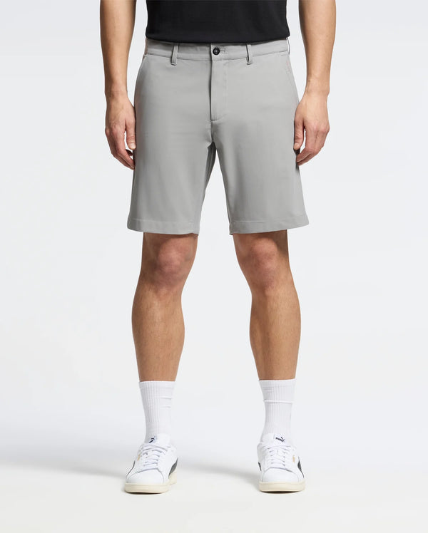 A person wearing Psycho Bunny's MENS 10K STEP SHORT (B6R969F200) stands facing forward in light gray, breathable shorts that reach just above the knees, paired with a black shirt, white sneakers, and white socks against a plain white background.