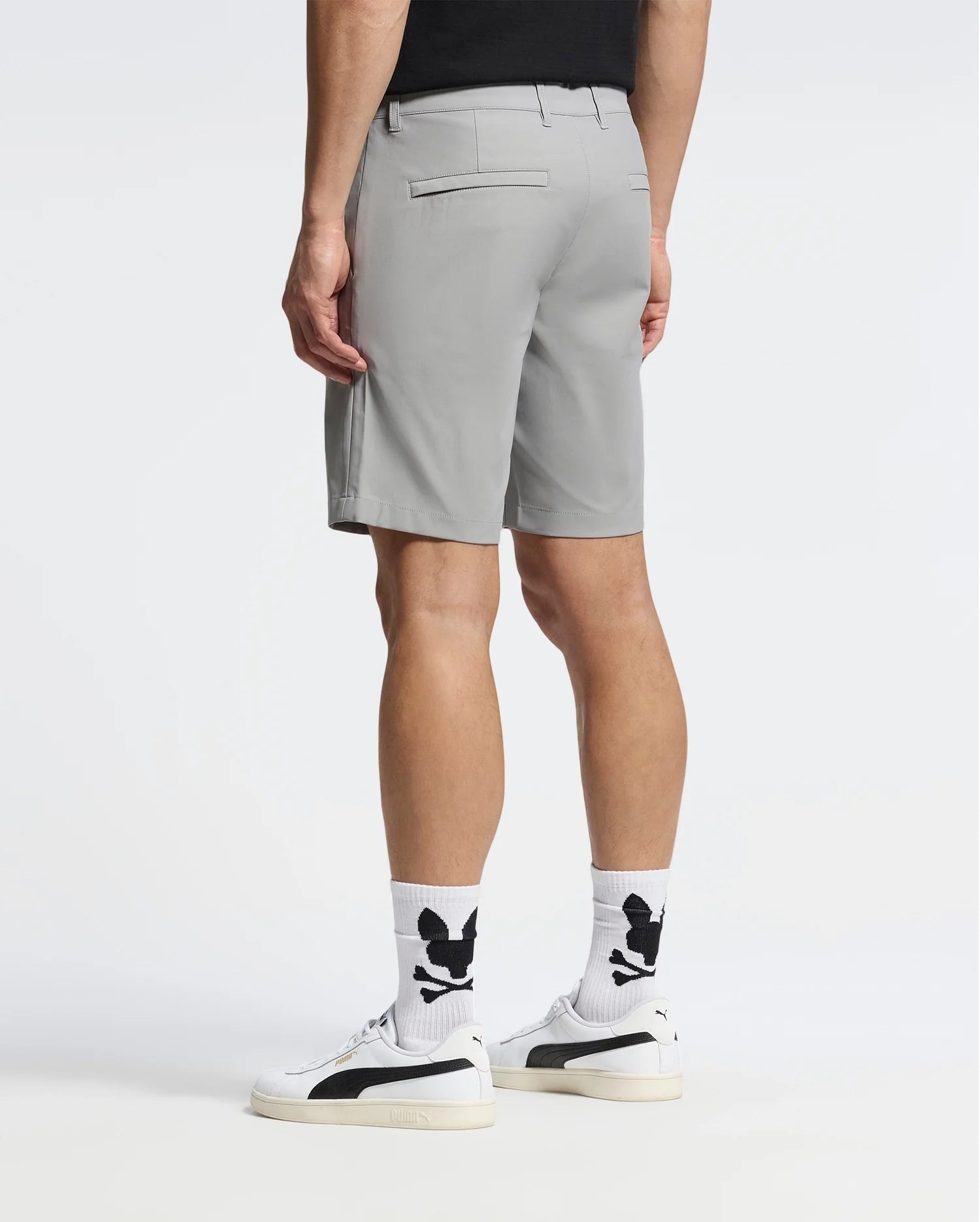 Dressed in light gray Psycho Bunny MENS 10K STEP SHORT - B6R969F200, white socks with black designs, a black shirt, and white sneakers with black stripes, the person stands facing away against a plain white background.