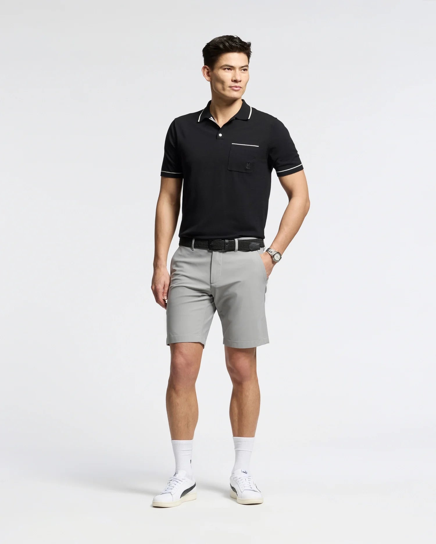 A person in a black polo and gray Psychobunny MENS 10K STEP SHORT - B6R969F200 stands smiling with one hand in their pocket. They wear white sneakers, socks, and a watch. The plain white background emphasizes the shorts' athletic breathability.