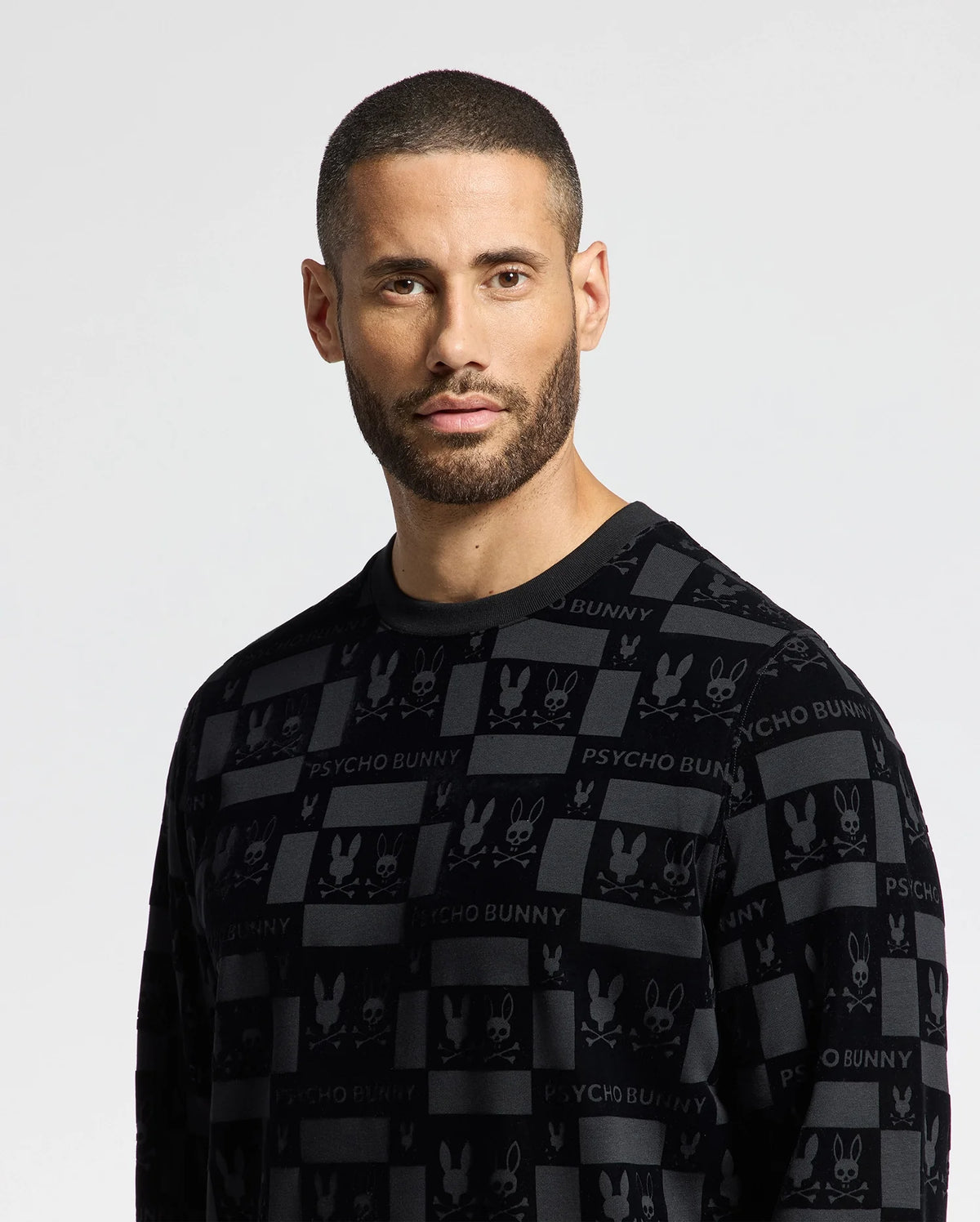 A man with short hair and a beard is wearing the stylish MENS ROMEO LIGHTWEIGHT SWEATSHIRT - B6S129E200 by Psycho Bunny. The black crew sweatshirt features "Psycho Bunny" and bunny logos, as he gazes directly at the camera against a neutral background, exuding confident simplicity.