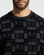 A person wearing the MENS ROMEO LIGHTWEIGHT SWEATSHIRT by Psycho Bunny, which is a stylish black crew sweatshirt adorned with the signature bunny skull logo and "PSYCHO BUNNY" wording. The person's face is partially visible, complemented by noticeable facial hair that enhances the edgy aesthetic.