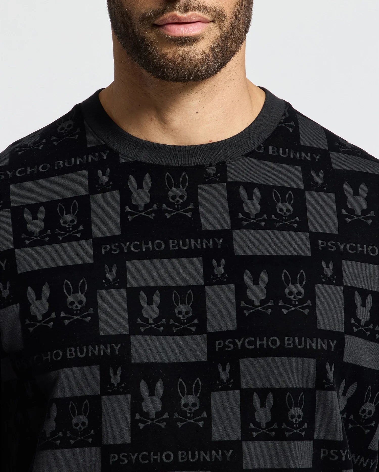 A person wearing the MENS ROMEO LIGHTWEIGHT SWEATSHIRT by Psycho Bunny, which is a stylish black crew sweatshirt adorned with the signature bunny skull logo and "PSYCHO BUNNY" wording. The person's face is partially visible, complemented by noticeable facial hair that enhances the edgy aesthetic.