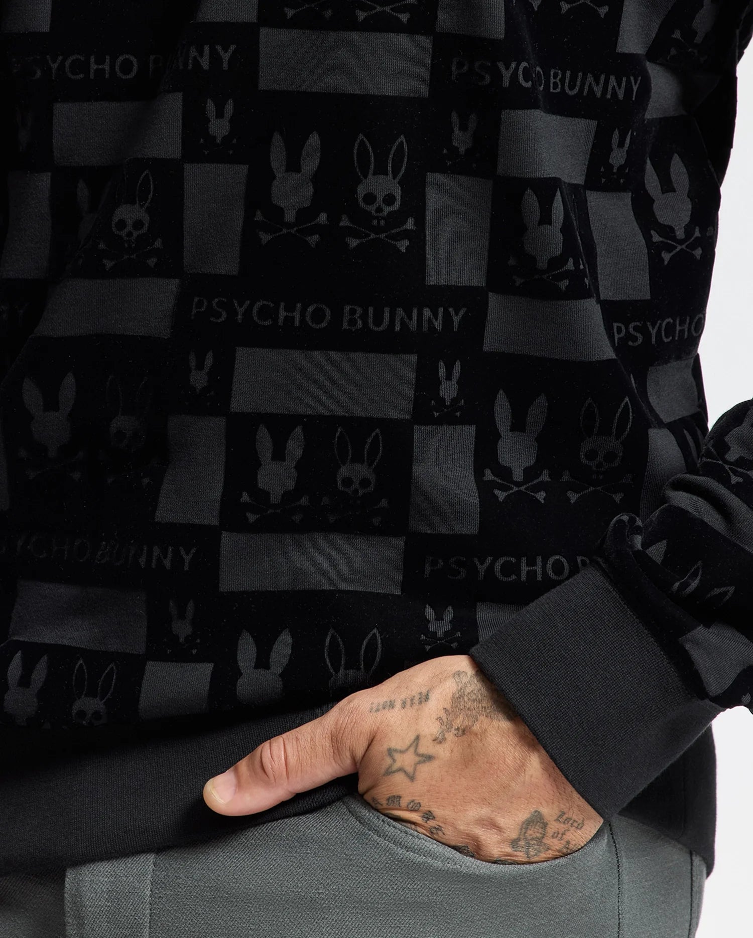 A person is wearing the MENS ROMEO LIGHTWEIGHT SWEATSHIRT by Psycho Bunny, a stylish black crew sweatshirt featuring bunny and skull designs along with the bold "PSYCHO BUNNY" text. Their hand, embellished with star tattoos and other designs, rests casually in their pocket, perfectly completing this effortlessly cool look.