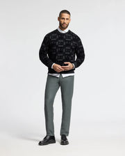 A man stands against a plain background, wearing a stylish black MENS ROMEO LIGHTWEIGHT SWEATSHIRT by Psycho Bunny, paired with a white collared shirt, gray pants, and black shoes. He is posing with his hands together and looking directly at the camera.