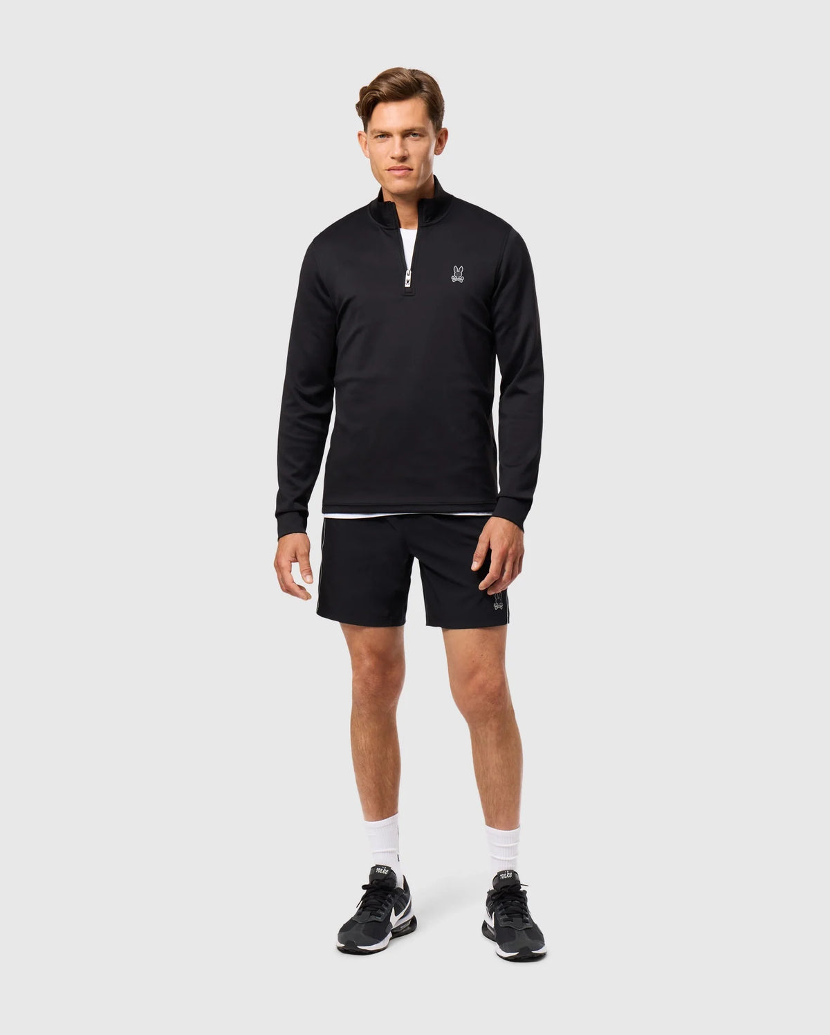 A man stands against a plain background, embodying the athleisure aesthetic. He wears a black long-sleeve Psycho Bunny LAURIER 1/4 ZIP HOODIE - B6S149D200, black shorts, white socks, and black sneakers. His hands are at his sides as he looks directly ahead.