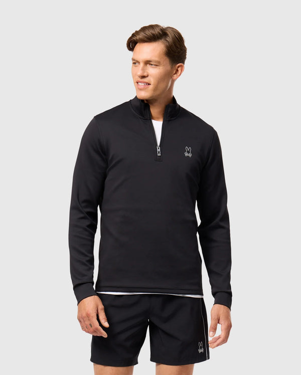 A man wearing a black Psycho Bunny Laurier 1/4 zip hoodie and black shorts stands against a plain white background. The cotton jersey hoodie, featuring a small embroidered logo on the chest, complements his athleisure aesthetic. He looks to the side with a relaxed expression.