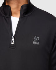 A close-up of a person wearing the Psycho Bunny MENS LAURIER 1/4 ZIP HOODIE - B6S149D200 in black with a white t-shirt underneath. Emphasizing an athleisure aesthetic, the hoodie features a rabbit skull and crossbones logo on the chest, with the zipper pull also adorned by a small rabbit logo.