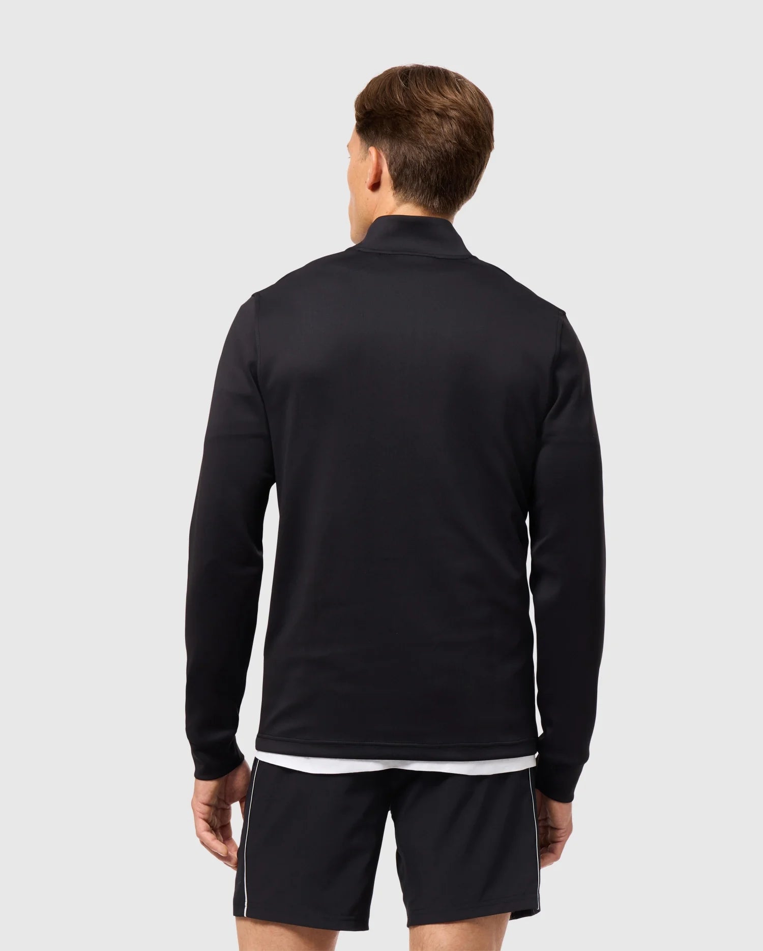 A person stands with their back to the camera, exuding an athleisure vibe in a black MENS LAURIER 1/4 ZIP HOODIE by Psycho Bunny, paired with black shorts featuring white trim, set against a plain light gray background.