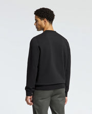 A person with short curly hair stands facing away from the camera, wearing a trendy Psycho Bunny MENS LAVETA French Terry Sweatshirt - B6S159E200 in black and gray pants against a plain light gray background.