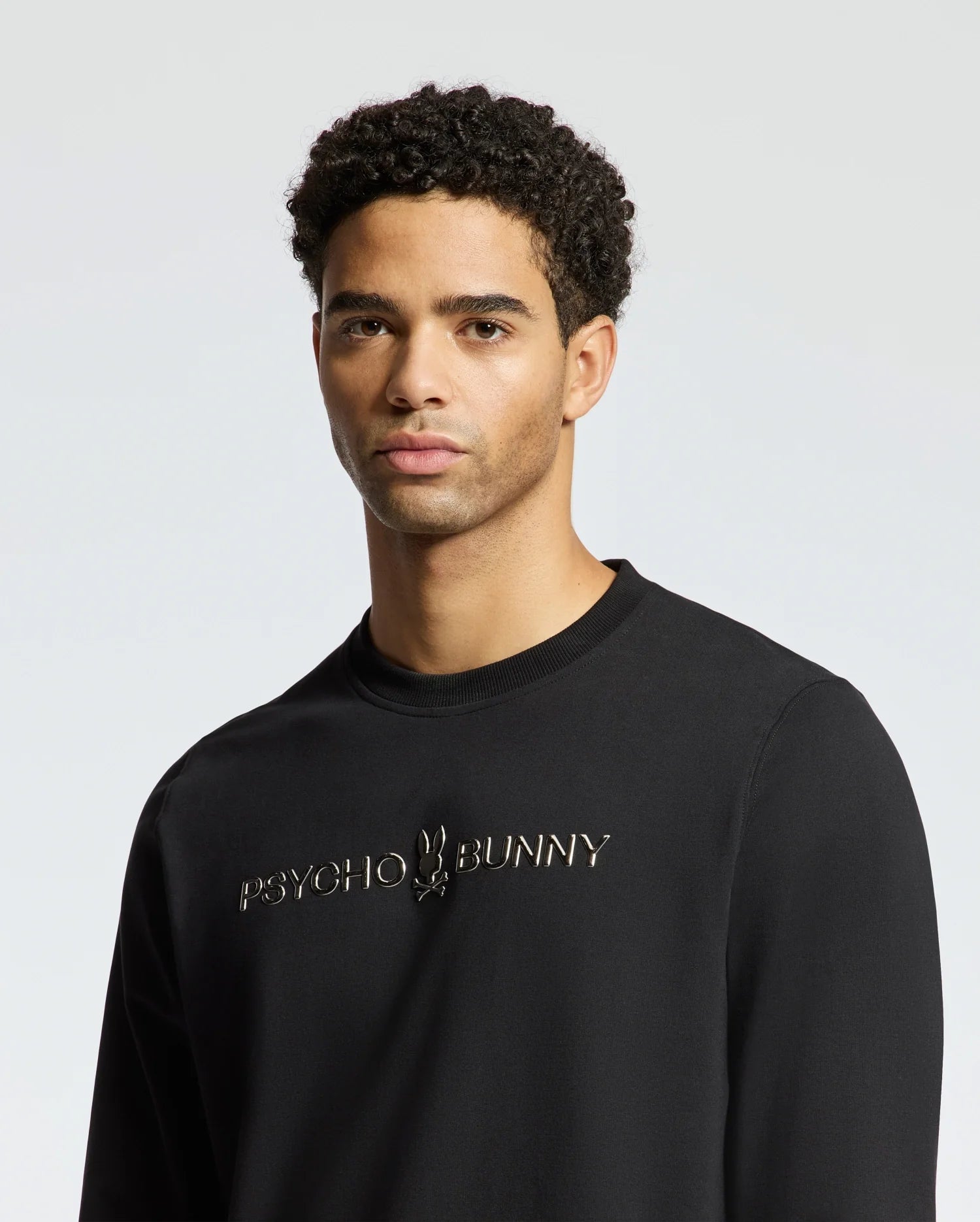 A person wearing the stylish black MENS LAVETA FRENCH TERRY SWEATSHIRT - B6S159E200 from Psycho Bunny stands against a plain, light background. This cozy cotton crew sweatshirt showcases the brand logo and name on the front. The individual has short curly hair and a neutral expression.