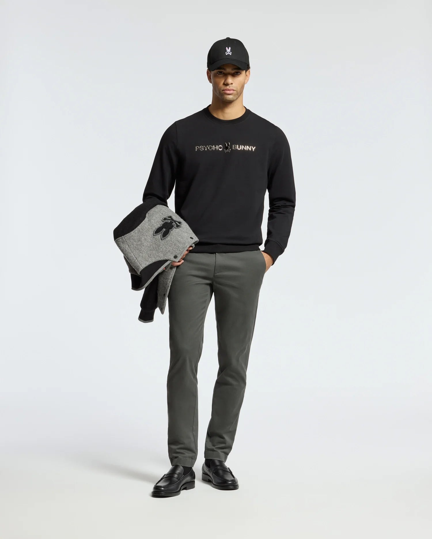 A person poses against a plain background, sporting the MENS LAVETA FRENCH TERRY SWEATSHIRT - B6S159E200 by Psycho Bunny in black, along with gray pants and black shoes. They hold a gray and black jacket and wear a black cap featuring the distinctive bunny logo.